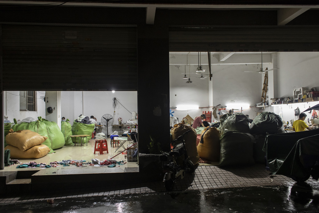 Fast Fashion 21 Blick in eine kleine Näh-Fabrik von der Straße aus. Es ist dunkel, der Raum ist hell erleuchtet.