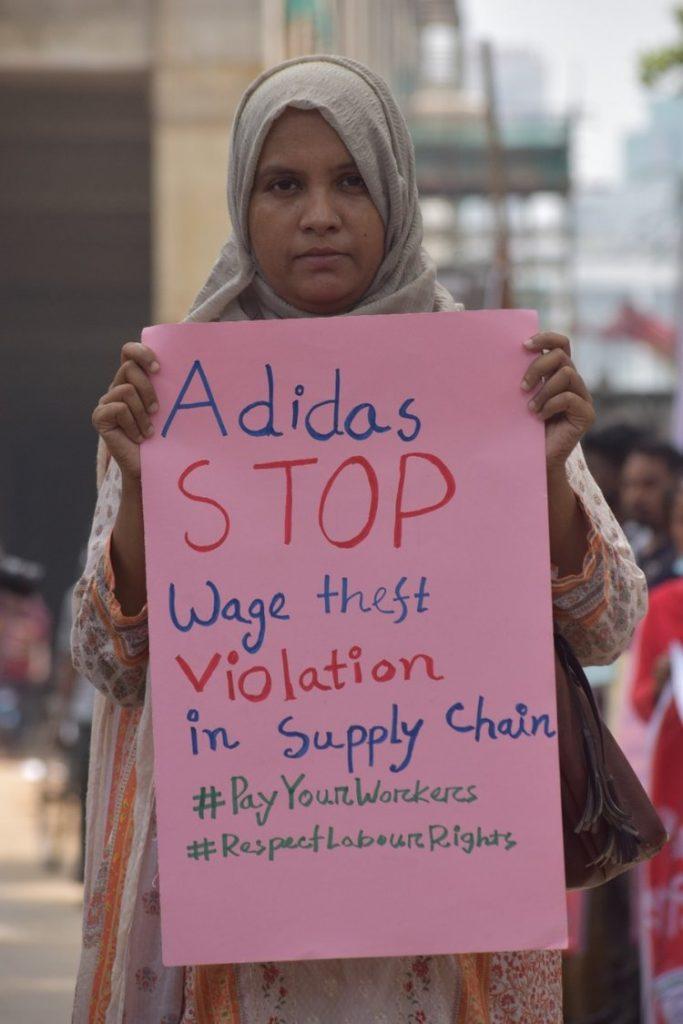 adidas – jetzt aktiv werden 1 Eine Frau hält ein rosafarbenes Plakat hoch. Daruaf steht unter anderem: "Adidas, Stop Wage theft"