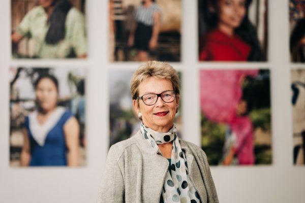 Eine Frau mit kurzen Haaren und Brille steht vor großen Fotografien von Frauen.