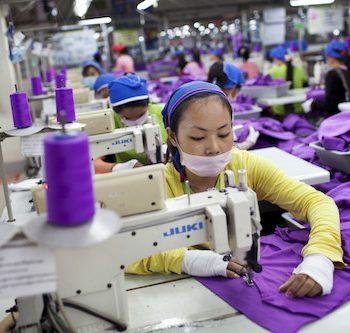 Eine Frau sitzt an einer Nähmaschine und näht lilafarbenen Stoff. Im Hintergrund sind viele weitere Näherinnen an ihren Maschinen zu sehen,