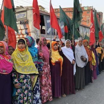 Eine Gruppe Frauen in bunten Saris steht am Straßenrand. Sie halten rote Fahnen.