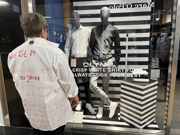 #yourturnOLYMP: Aktionen in Berlin 2 Eine Frau steht mit dem Rücken zum/zur Betrachter*in vor dem Schaufenster eines Olymp-Ladens. Sie trägt ein weißes Hemd, auf das mit rotem Stift geschrieben wurde.