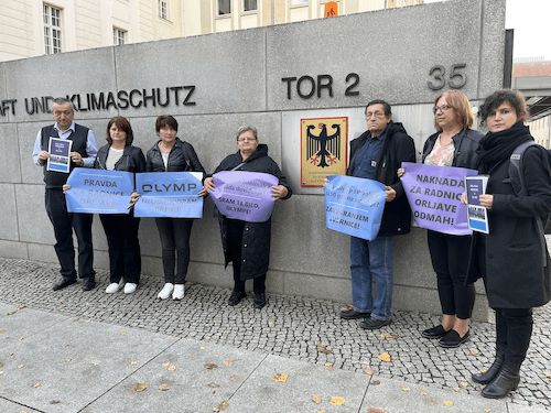 Sieben Personen stehen vor einer grauen Mauer. Sie halten blaue und lilafarbene Plakate in den Händen.