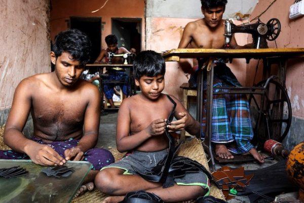 Vision Mission Kampagne für Saubere Kleidung Ein Kind und ein junger Mann sitzen auf dem Boden und nähen mit der Hand Leder. Im Hintergrund arbeiten zwei Männer an Nähmaschinen.