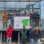 Eine Gruppe Menschen steht vor einem Plakat. Sie tragen Jacken.