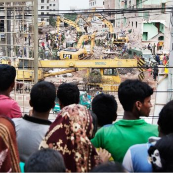 Elf Jahre nach Rana Plaza: Textilfabriken sicherer, Auslöser der Katastrophe bleiben 1 Eine Gruppe Menschen - von hinten zu sehen - stehen vor einem Gitter. Dahinter sind die Trümmer eines eingestürzten Gebäudes zu sehen.