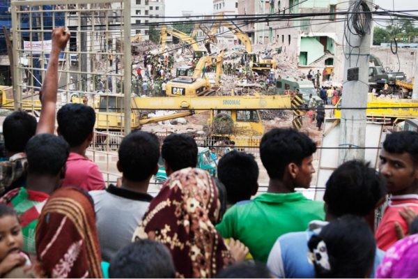 Elf Jahre nach Rana Plaza: Textilfabriken sicherer, Auslöser der Katastrophe bleiben 50 Eine Gruppe Menschen - von hinten zu sehen - stehen vor einem Gitter. Dahinter sind die Trümmer eines eingestürzten Gebäudes zu sehen.