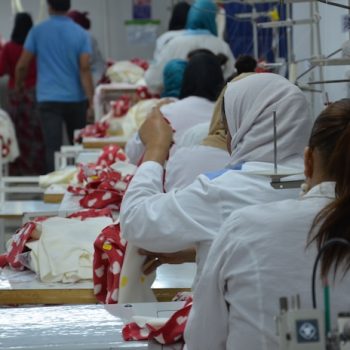 Frauen sitzen hintereinander an Nähtischen. Sie sind von hinten zu sehen.