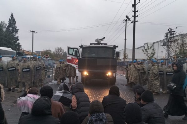 Soldaten und ein Militär-Lkw stehen einer Gruppe Menschen gegenüber. Der Himmel ist grau.