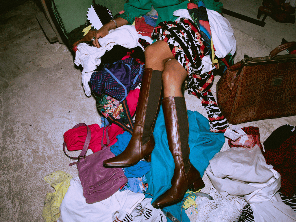 Fast Fashion 19 Eine Person sitzt auf einem Haufen Kleidung, man sieht nur ihre Beine