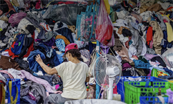 One-Earth Fashion: 33 Ziele für ein gerechtes Modesystem 18 Eine Frau mit roter Mütze steht vor einem Berg Kleidung. Vor ihr steht ein Ventilator.