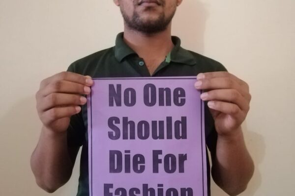 Bangladesch: Erneut schwere Unfälle in Textilfabriken 2 Ein junger Mann hält ein lilafarbenes Schild in die Kamera. Darauf steht: "No One Should Die For Fashion"